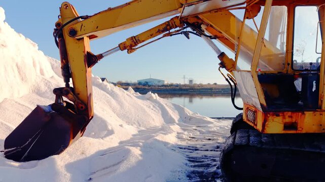 Excavator arm over salt stockpile at coastal salt works, Bulgaria. Low-angle close view of yellow rusty boom and bucket framing a white salt mound, reflective pond and blue industrial building behind.
