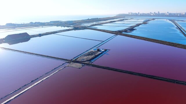 Aerial view of salt pans and harvest equipment at sunrise, Bulgaria. Elevated angle of excavator on central island surrounded by pink and blue reflective ponds, rail tracks, hazy sky.