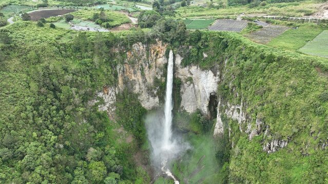 Aerial view of Sipiso Piso Waterfall plunging down a steep rocky cliff surrounded by lush green forests and agricultural fields in Merek, North Sumatra, Indonesia.
