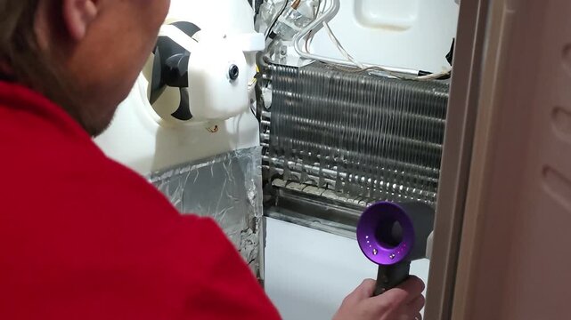 Repairman using hair dryer for defrosting refrigerator evaporator coil. Technician removing ice buildup from freezer cooling element. Appliance maintenance service.