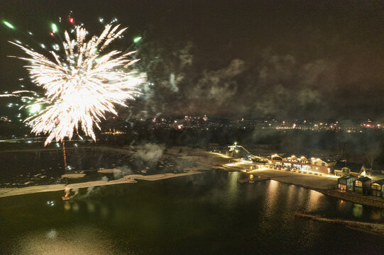 Aerial view of fireworks exploding over a lake at night with illuminated buildings and a beach area on the shore in Stary Sacz, Lesser Poland Voivodeship, Poland.