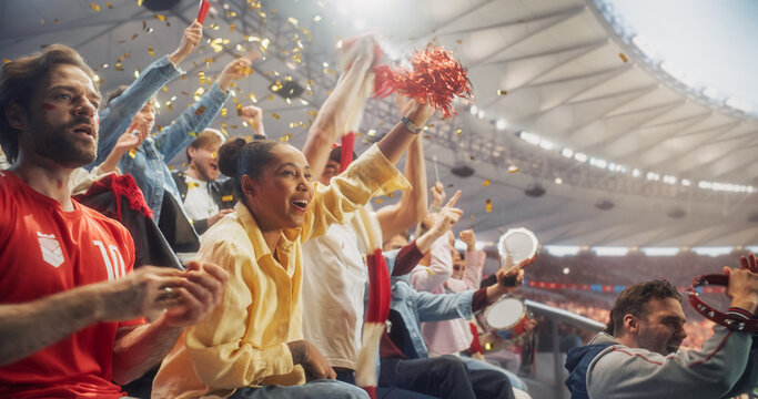 Diverse Men and Women in Stadium Seats Roar With Relieved Joy After a Decisive Soccer Championship Moment, as a Woman Lifts a Red Pompom, Fans Beat Tambourines and Gold Confetti Swirls Above.