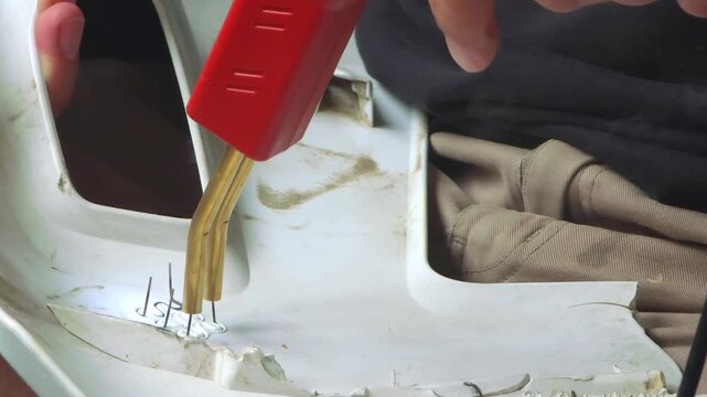 Technician repairing cracked white plastic car bumper with hot stapler tool. Man fusing broken vehicle body part using metal staples for automotive restoration.