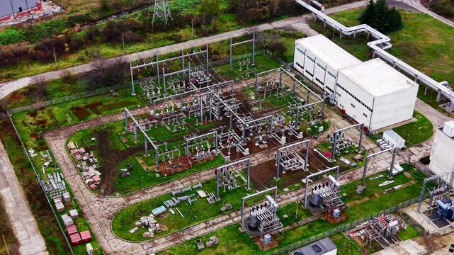 Overhead aerial view of electrical substation in industrial facility, Bulgaria. Power distribution equipment, transformers, metal pylons and pipelines on green grass.