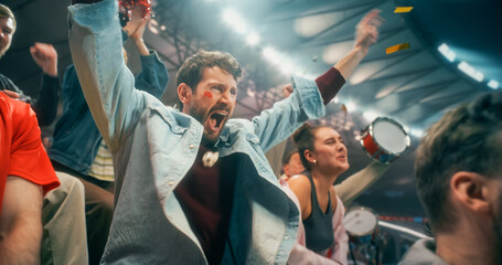 Fototapeta premium Close Portrait of White Male Soccer Football Supporter With Red White Face Paint, Arms Raised Amid Blurred Blue Streamers, Happy Cheer in Packed Championship Stadium Crowd During Cup League Match.