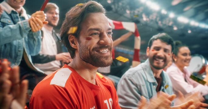 Close Portrait of White Male Soccer Football Supporter With Red and White Face Paint, Smiling in Packed Championship Stadium as Diverse Fans Wave Flags and Cheer During Intense Cup Tournament Match.
