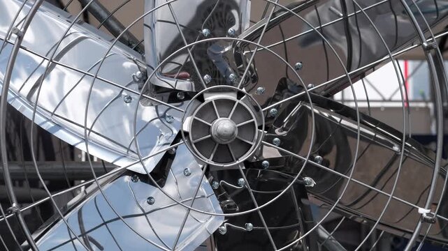 Tight shot of a polished chrome industrial fan viewed through a circular wire safety grille. Three reflective metal blades fill the frame with bright specular