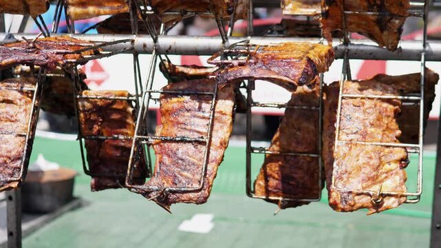 Close-up view of multiple marinated pork ribs suspended in wire grilling baskets from a horizontal metal rack above an outdoor grill. Charred, caramelized