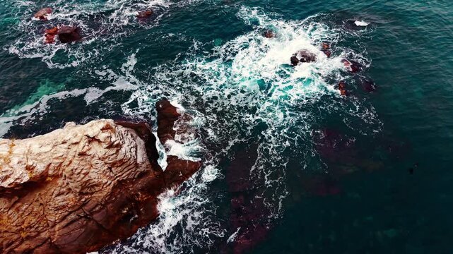 Directly overhead drone shot of white surf churning around stone formations in deep teal water. Sea waves breaking over dark coastal rocks, Black Sea coast