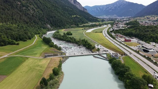 Drone footage flying across a hydroelectric dam with turquoise reservoir water upstream and water diversion infrastructure downstream. Highway transportation routes run alongside the energy facility.