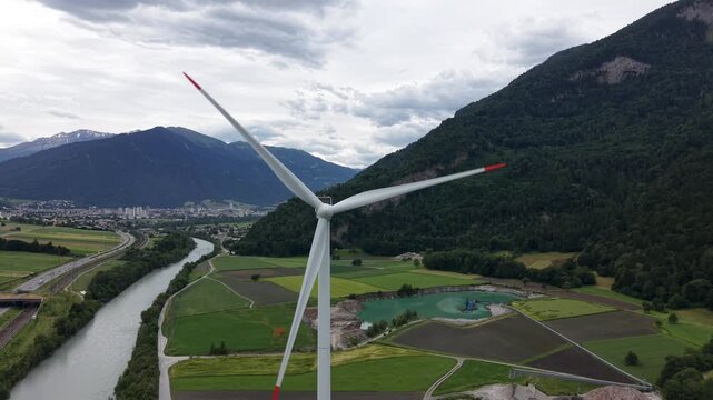 Aerial drone footage approaching a rotating wind turbine surrounded by mountains. Renewable energy and sustainable power generation concept.

