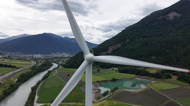 Static drone shot positioned near the center of a wind turbine rotor with blades rotating against a dramatic alpine mountain backdrop.