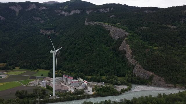 Aerial drone footage approaching a wind turbine located above a gravel processing facility near a river in a mountain valley environment
