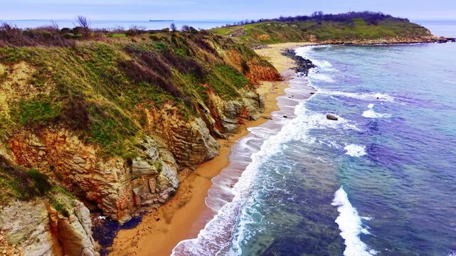 Panoramic drone perspective of empty coast meeting the ocean under a cloudy winter sky. High angle view of a long sandy shoreline below grassy cliffs.