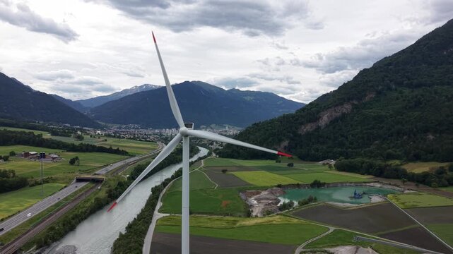 Drone footage of a wind turbine surrounded by mountains, with a passing train, highway and river visible below. Renewable energy and modern infrastructure concept.
