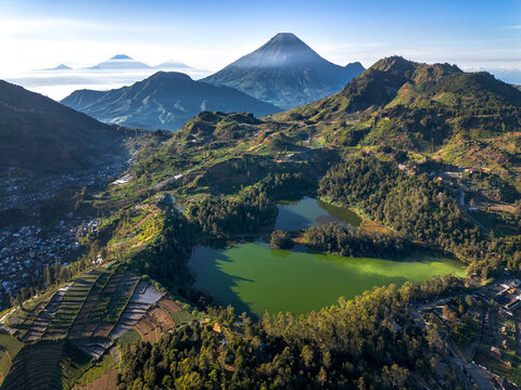 Aerial view of the green volcanic lake Telaga Warna surrounded by terraced fields and dense forests with Mount Sindoro in the background under a clear sky in Indonesia.