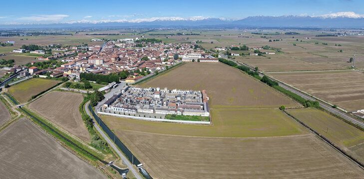 Aerial view of the town surrounded by vast agricultural fields and a cemetery with snow-capped mountains in the background San Germano Vercellese, Piedmont, Italy.