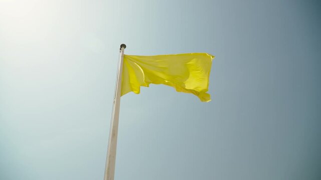 Yellow flag on flagpole against sunny blue sky on beach, wide low angle shot. Indicator of wave danger for swimming on sea coastline without people waving in wind
