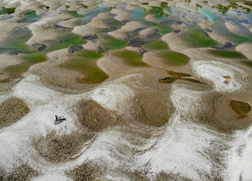 Aerial view of a person riding a bicycle across a dry riverbed with patterns of water pools and sandy mounds in Bogura, Bangladesh.