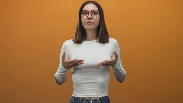Woman showing palms forward, hands visible and wearing glasses in studio with an orange wall; calm reflection.