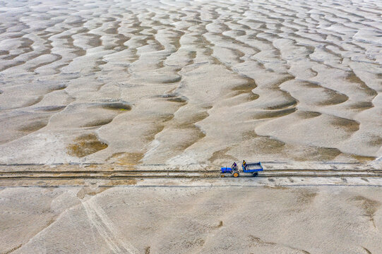 Aerial view of a blue tractor with two people on board driving on a sandy track across a vast riverbed with unique ripple patterns and sand dunes Bogura, Bangladesh.