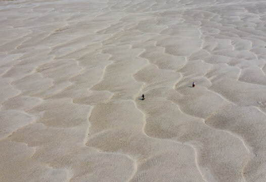 Aerial view of two people walking across a vast rippled sandbar with wavy textures and soft light in Bogura, Bangladesh.