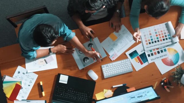 Top view of manager writing idea while team standing and giving comment. Professional designer design logo while looking at color palette and writing idea down in paper with color marker. Symposium.