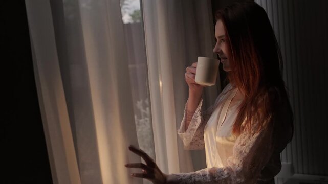 Young redhead woman in a satin robe holding a cup of tea or coffee near a window at home. Cozy evening atmosphere, relaxation, mindfulness and self care lifestyle concept