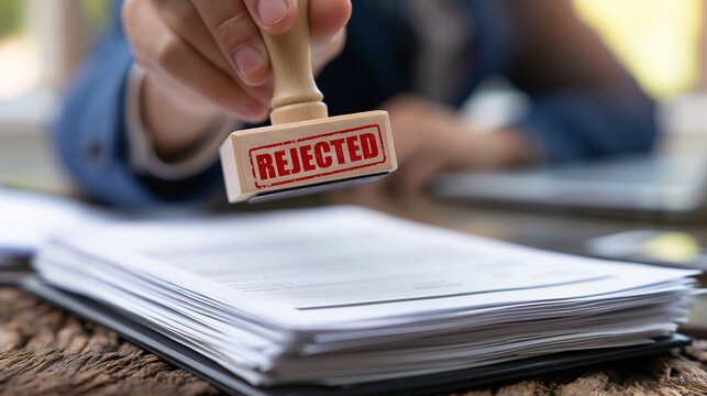Rejected Document Stamp: A hand holds a stamp with the word REJECTED prominently displayed, about to mark a stack of important documents, symbolizing denial and disapproval in a formal setting.