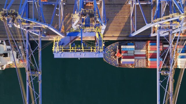 Stunning top-down aerial perspective of a massive cargo ship docked at a commercial port while industrial gantry cranes efficiently load and unload colorful shipping containers from the vessel