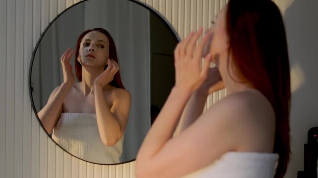 Young redhead woman wearing a towel and hydrating sheet mask standing in front of a mirror during an evening self care routine at home. Beauty, wellness and skincare concept
