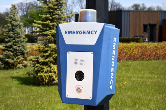 Emergency call box with camera and flashing light mounted on a pole beside a grassy park and campus building. A blue alarm station provides safety and emergency communication