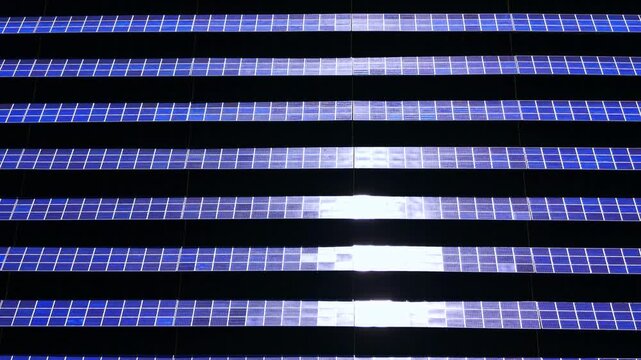 Symmetric overhead perspective of alternative energy array with central sun glint reflection on blue modules. Horizontal rows of solar panels at photovoltaic farm.