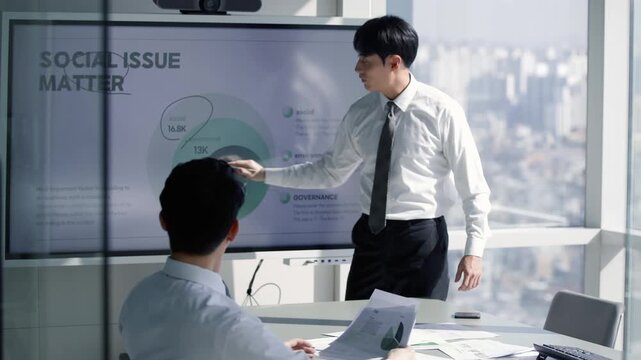 Asian businessman presenting ESG report on a large digital screen in a modern office, discussing social issue matters with a colleague during a corporate meeting.