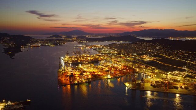 Stunning aerial footage capturing the illuminated container terminal at the commercial port of piraeus in greece, showcasing the bustling international trade hub against a beautiful sunset sky