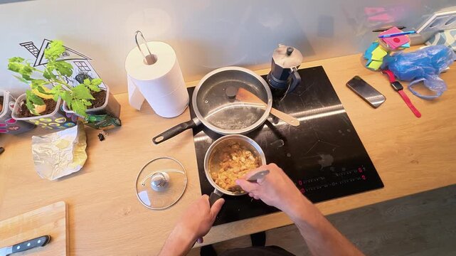 POV overhead shot of a person stirring food in a saucepan on an induction cooktop, surrounded by kitchen tools, fresh herbs, and vibrant countertop items under natural light. (197 characters)