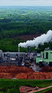Industrial high-angle shot over the Stora Enso sawmill in Latvia. Features thick white steam venting from a factory smokestack next to large piles of processed woodchips and a green forest line.