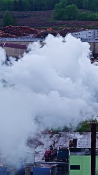 Industrial high-angle shot over the Stora Enso sawmill in Latvia. Features thick white steam venting from a factory smokestack next to large piles of processed woodchips and a green forest line.