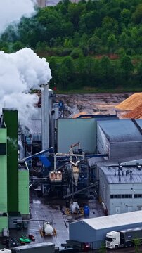 Industrial high-angle shot over the Stora Enso sawmill in Latvia. Features thick white steam venting from a factory smokestack next to large piles of processed woodchips and a green forest line.
