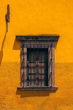 Sunlit yellow wall with aged window. Black iron bars frame a wooden window. Weathered stone trim casts soft shadows. Rustic charm hints at old world character. Ideal for rustic, vintage