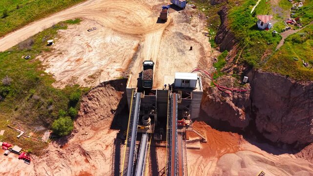 Commercial vehicle tipping raw minerals into a crushing hopper next to long conveyor structures. Dump truck unloading crushed stone at processing plant.