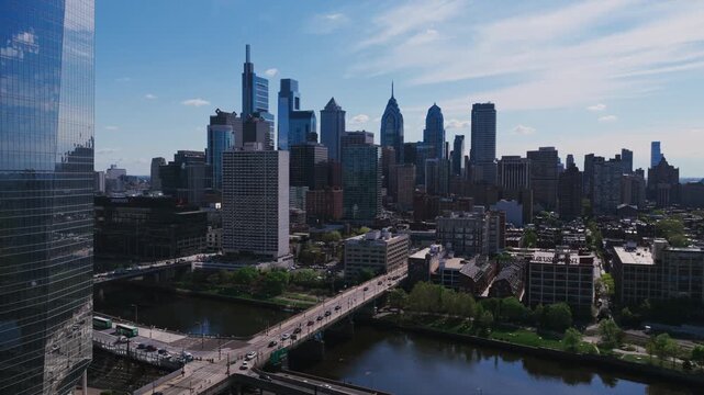 Aerial drone view of Philadelphia's full Center City skyline reflected in a glass tower, with the Schuylkill River, a bridge, and Comcast buildings prominent.