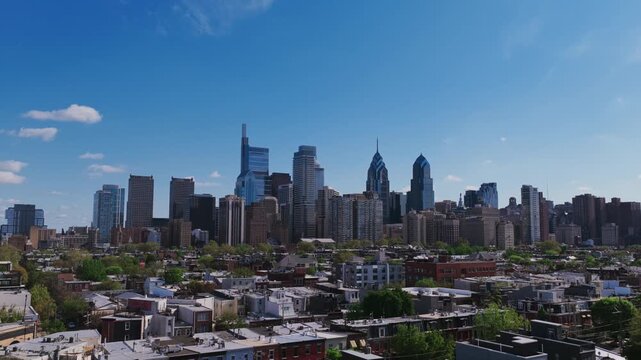Aerial drone view of Philadelphia's iconic skyline featuring Comcast towers and One Liberty Place rising above rowhouse neighborhoods under a bright blue sky.