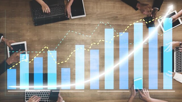 A business team engages in collaborative performance analysis using digital devices while reviewing growth graphs and statistics on a wooden desk in an office setting. Copula
