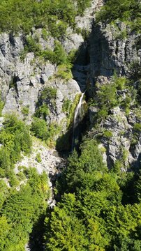 Aerial drone footage slowly flying toward Grunas Waterfall in Theth National Park, Albania. The camera approaches the waterfall from above the forest canopy, revealing the dramatic rocky cliffs and lu