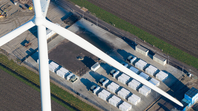 Aerial view of a wind turbine and a battery energy storage system with rows of white containers on a concrete pad near agricultural fields in Dronten, Flevoland, Netherlands.