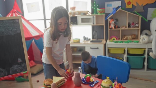 Girl placing toy sandwich with hands on a small table while a boy kneels beside toy kitchen and chalkboard in a playroom building; playful focus.