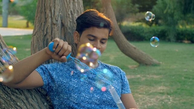 A cheerful youngster having fun while launching soap bubbles sitting in the park. Closeup shot of an Indian male wearing a blue T-shirt happily spending his free - remembering childhood and relaxing