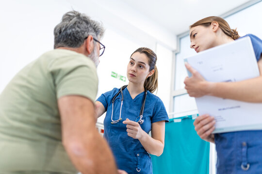Female medical doctors consulting senior male patient in clinic, healthcare concept. Two professional nurses listening to elderly man case history during clinical examination in hospital ward room.