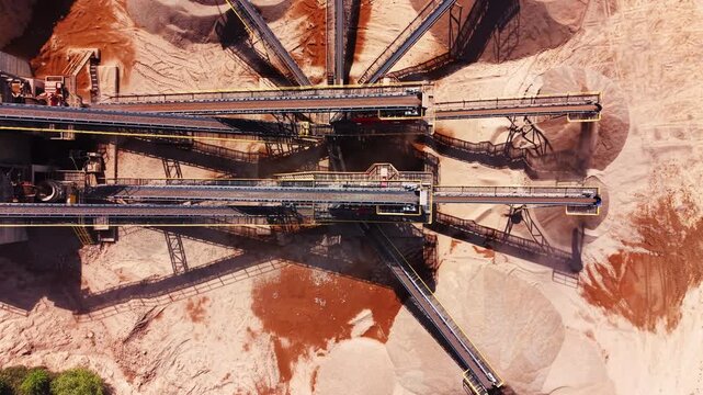 Double industrial belts transporting crushed rock to sand stockpiles below. Top-down aerial view of parallel aggregate conveyor belts.